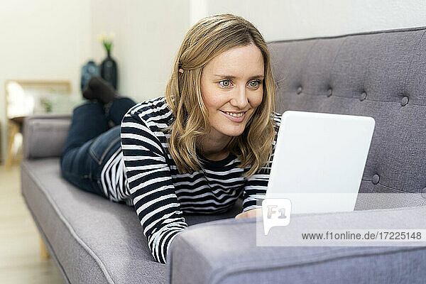 Lächelnde Frau  die ein digitales Tablet benutzt  während sie zu Hause auf dem Sofa liegt