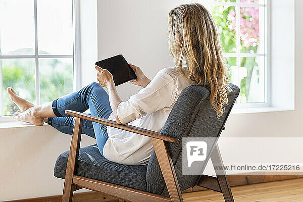 Frau mit blondem Haar  die ein digitales Tablet hält  während sie zu Hause auf einem Sessel sitzt