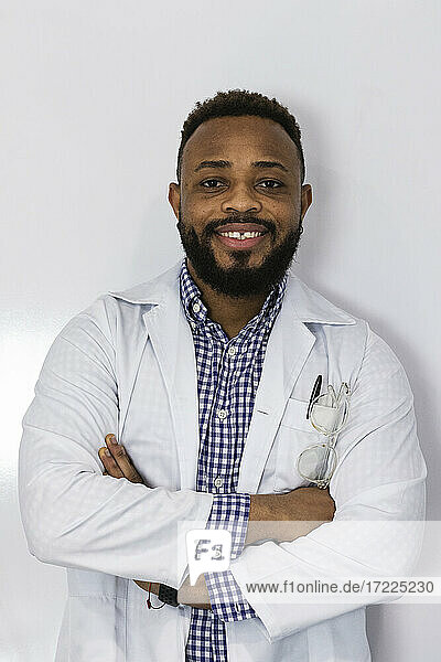 Lächelnder junger Arzt steht mit verschränkten Armen vor einer weißen Wand