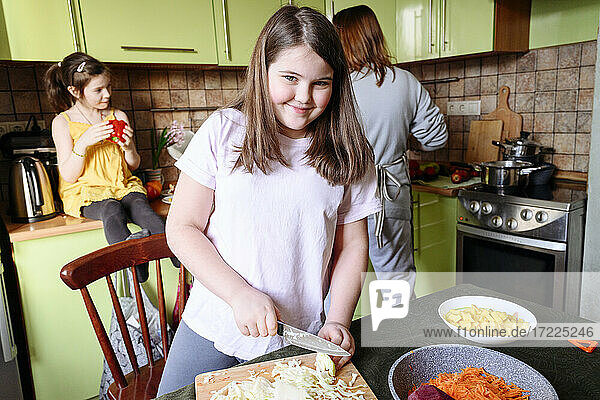 Lächelndes Mädchen  das Kohl schneidet  während Mutter und Schwester im Hintergrund Essen zubereiten