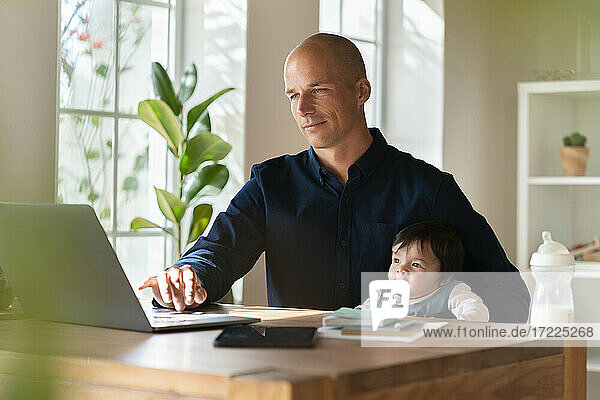 Männlicher Unternehmer arbeitet am Laptop  während er sein Baby im Home Office hält