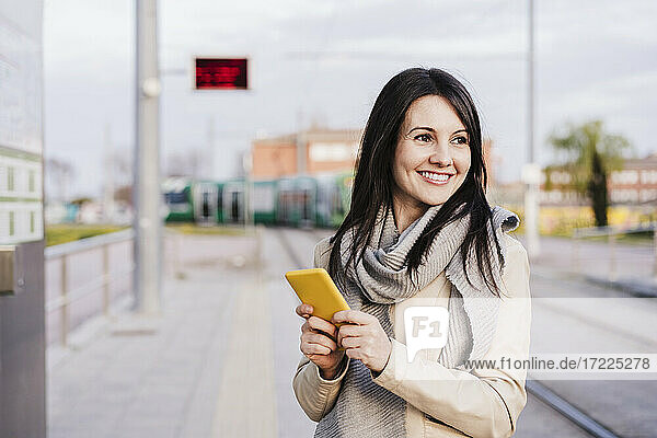 Smiling beautiful woman contemplating while standing with smart phone at railroad station