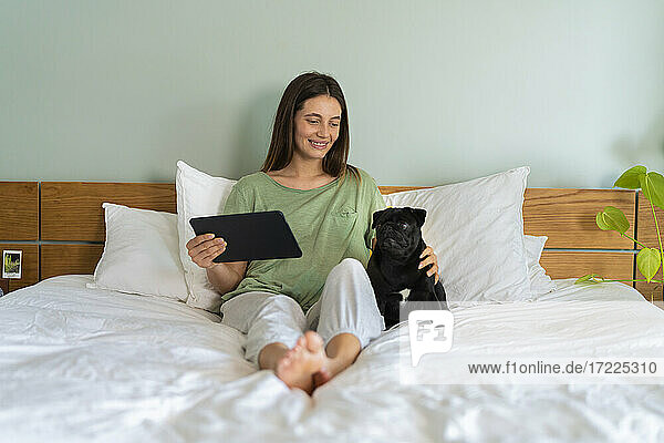 Young woman holding digital tablet sitting with Pug dog on bed at home