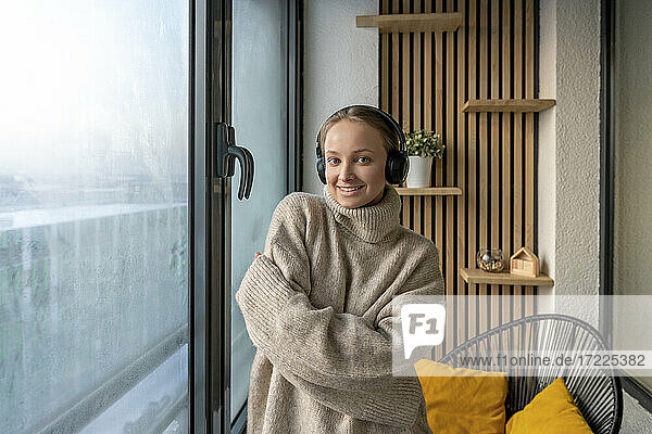 Junge lächelnde Frau mit Kopfhörern  die mit verschränkten Armen vor einem Fenster zu Hause steht