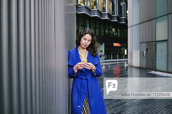 Geschäftsfrau in blauem Trenchcoat arbeitet an einem Mobiltelefon und lehnt sich an die Wand