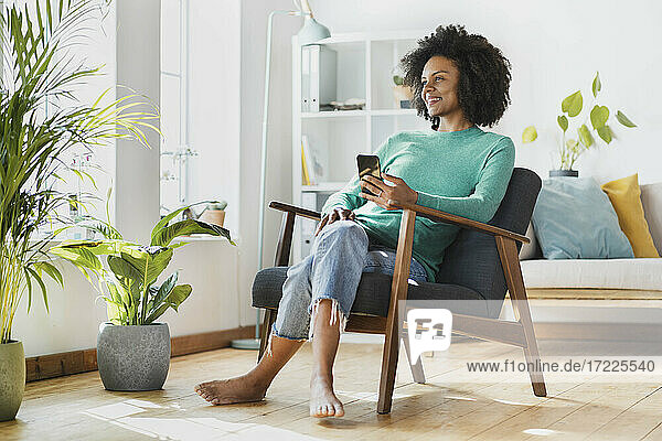 Lächelnde Frau  die ein Mobiltelefon benutzt  während sie zu Hause sitzt
