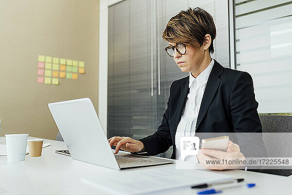 Geschäftsfrau  die einen Laptop benutzt und ein Mobiltelefon am Schreibtisch im Büro hält