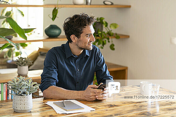 Smiling male freelance worker holding smartphone while looking away at home