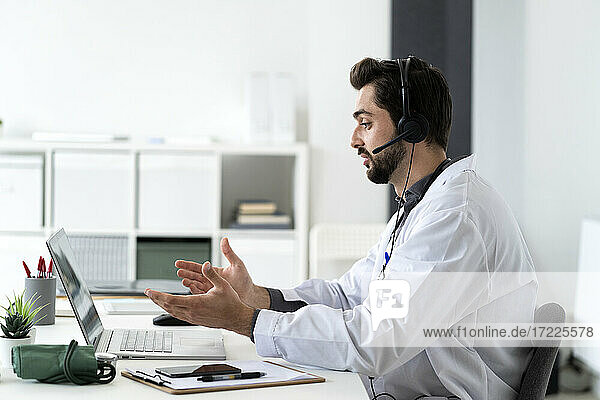 Männlicher Arzt trägt Kopfhörer  während er über einen Laptop eine Videokonferenz abhält
