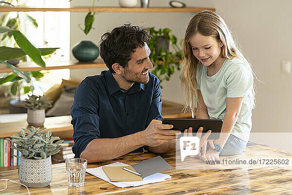 Happy father with digital tablet looking at daughter while discussing at home