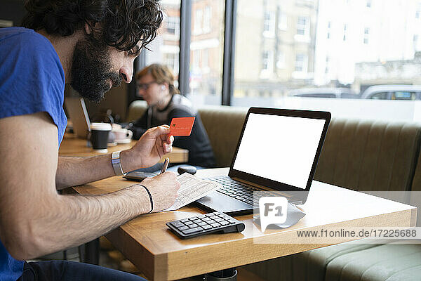 Männlicher Unternehmer  der einen Kredit in der Hand hält  während er eine Online-Zahlung am Laptop im Büro vornimmt
