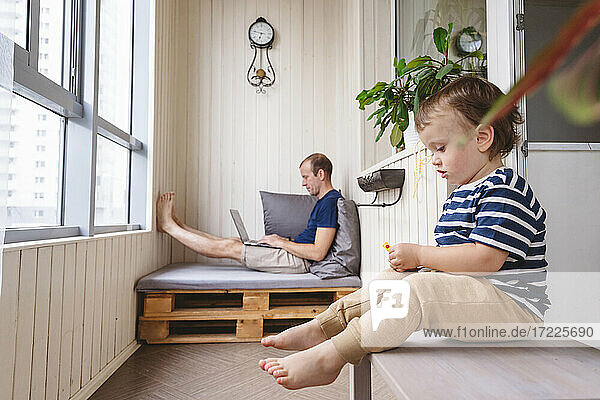 Verspieltes Kind sitzt auf dem Tisch  während der Vater im Büro zu Hause am Laptop arbeitet