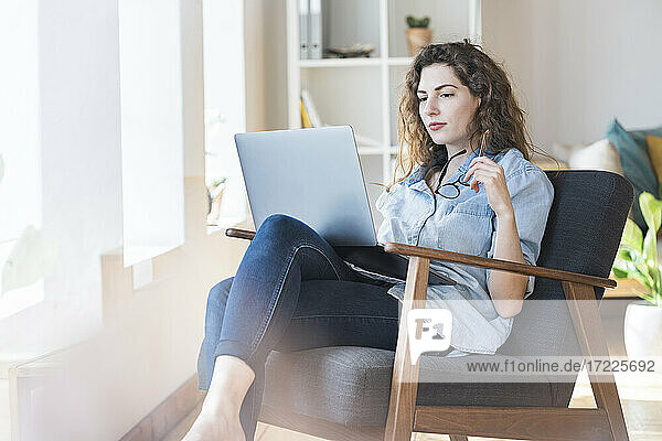 Nachdenkliche junge Frau hält eine Brille  während sie auf einem Stuhl zu Hause auf ihren Laptop schaut