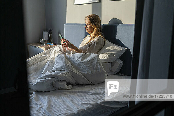 Frau  die ein Mobiltelefon benutzt  während sie mit einer Decke auf dem Bett zu Hause sitzt