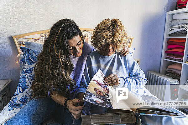 Lächelnde Tochter schaut sich ein Fotoalbum an  während sie mit ihrer Mutter im Schlafzimmer sitzt