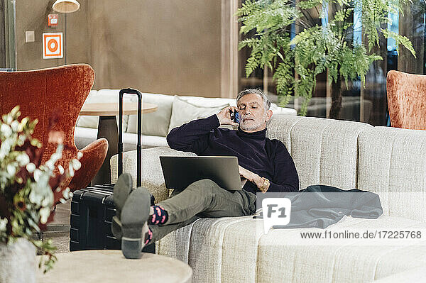 Geschäftsmann  der mit dem Handy telefoniert  während er sich mit seinem Laptop auf dem Sofa im Hotel ausruht