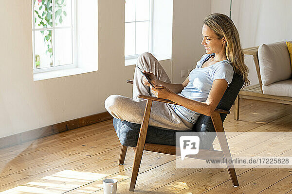 Lächelnde Frau  die ein digitales Tablet benutzt  während sie auf einem Stuhl im Wohnzimmer sitzt