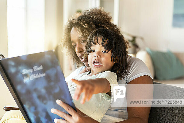 Kleinkindes Mädchen gestikuliert  während die Mutter ihr im Wohnzimmer ein Buch vorliest