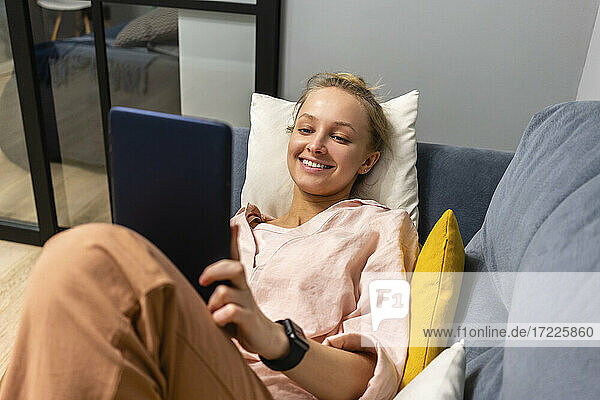 Lächelnde junge Frau  die ein digitales Tablet benutzt  während sie zu Hause auf dem Sofa liegt
