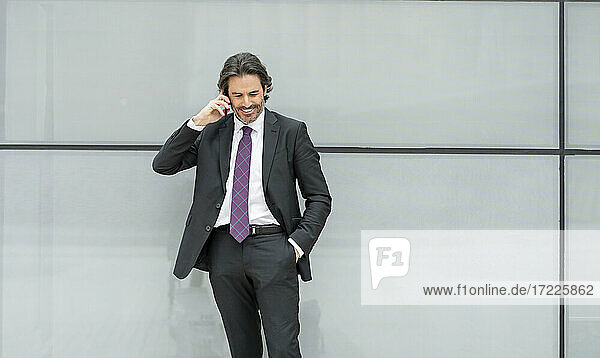 Lächelnder Geschäftsmann mit Hand in der Tasche  der vor einer weißen Wand mit einem Mobiltelefon spricht