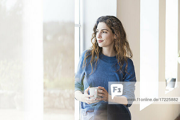 Junge Frau hält Kaffeetasse  während sie am Fenster im Wohnzimmer sitzt