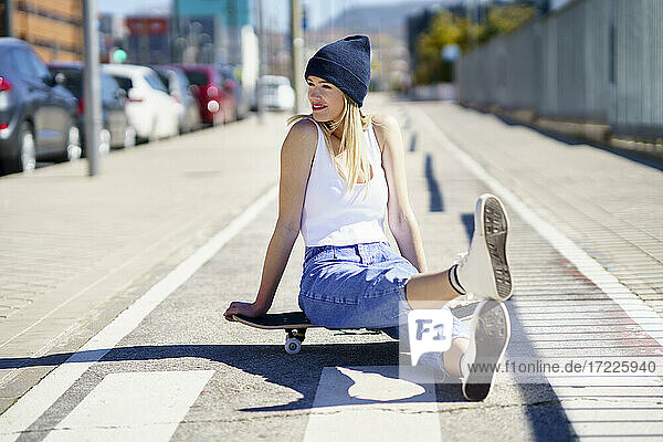 Smiling blond woman looking away while sitting on footpath during sunny day