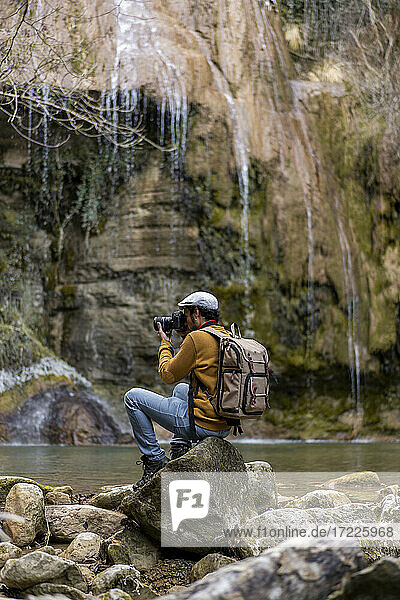 Männlicher Wanderer  der auf einem Felsen am See im Wald sitzt und fotografiert