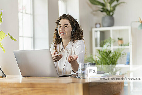 Lächelnde Frau gestikuliert  während sie im Büro zu Hause über Kopfhörer spricht