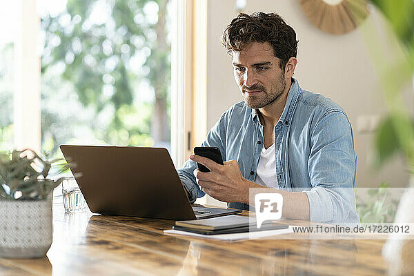 Seriöser männlicher Freiberufler  der ein Mobiltelefon hält und einen Laptop am Tisch benutzt