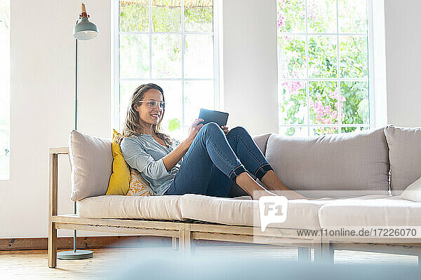 Smiling woman with eyeglasses holding digital tablet on couch in living room