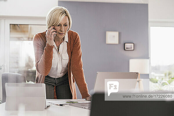 Weibliche Fachkraft  die auf einen Laptop schaut  während sie über ein Mobiltelefon im Heimbüro diskutiert