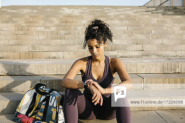 Weibliche Athletin  die die Zeit kontrolliert  während sie mit einer Tasche auf einer Treppe an einem sonnigen Tag sitzt