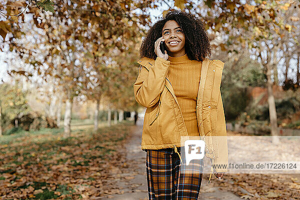 Smiling Afro woman talking on smart phone while walking on footpath during autumn