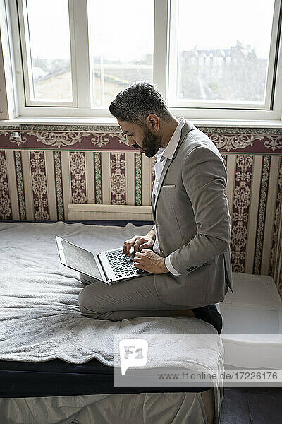 Männlicher Unternehmer  der einen Laptop benutzt  während er zu Hause auf dem Bett sitzt