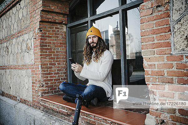 Hipster-Mann mit Smartphone auf dem Fensterbrett sitzend