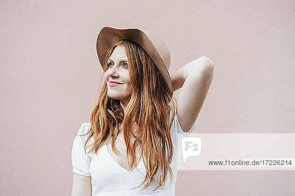 Smiling redhead woman standing in front of pink wall while looking away