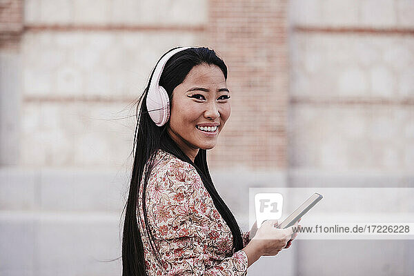Lächelnde Frau mit Kopfhörern  die ihr Smartphone an die Wand hält