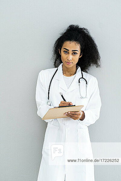 Confident female doctor holding clipboard while standing in front of wall