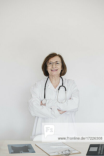 Lächelnde Ärztin mit verschränkten Armen vor einem Schreibtisch im Krankenhaus stehend
