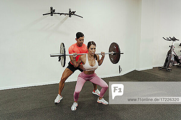 Fitness instructor guiding female athlete while exercising with barbel in gym