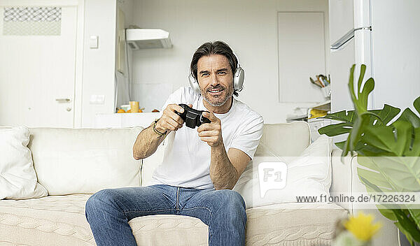 Aufgeregter Mann spielt ein Videospiel  während er auf dem Sofa im Wohnzimmer sitzt