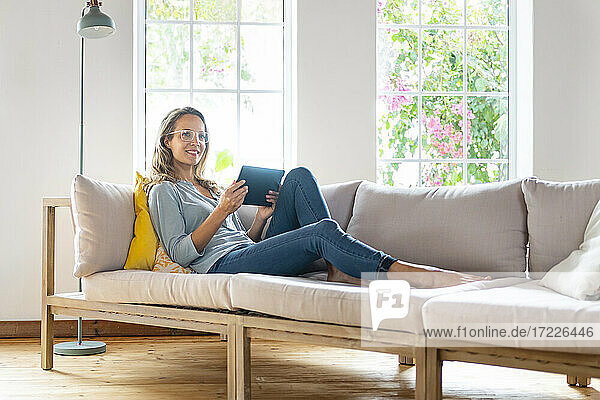 Smiling beautiful woman looking away while holding digital tablet on sofa in living room