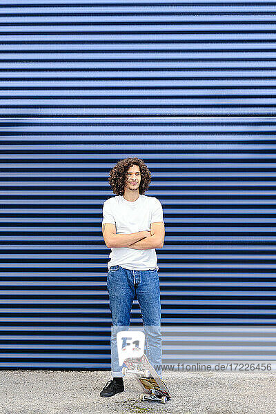 Kaukasischer junger Mann mit verschränkten Armen auf einem Skateboard vor einem blauen Fensterladen stehend