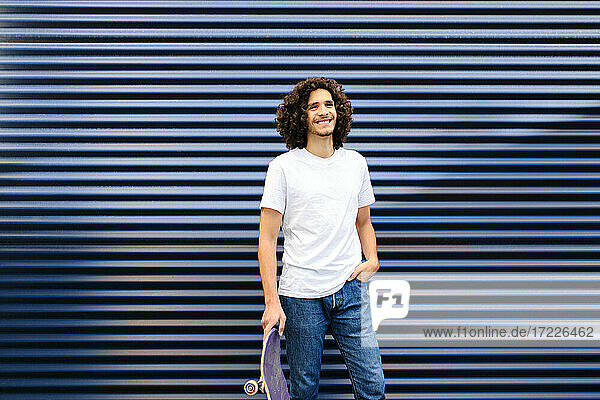 Junger Mann mit lockigem Haar  der ein Skateboard hält  während er vor einem blauen Fensterladen steht