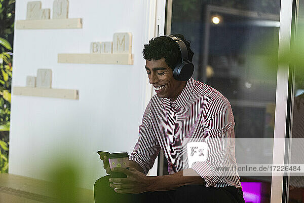 Lächelnder männlicher Fachmann mit Kopfhörern  der ein Smartphone benutzt  während er einen Kaffee im Büro trinkt