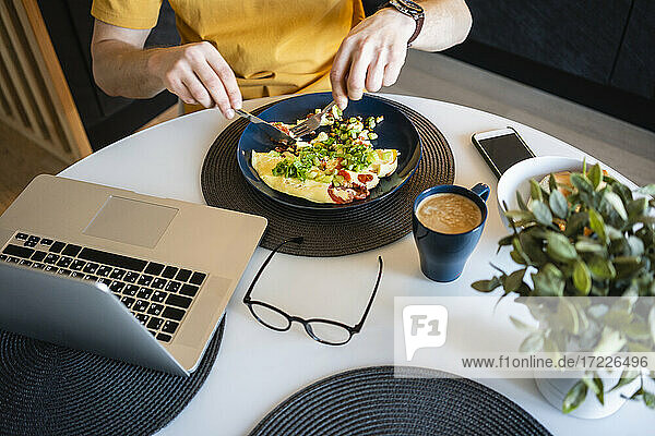 Geschäftsmann isst Omelett und Gemüse  während er mit seinem Laptop an einem Tisch im Haus sitzt