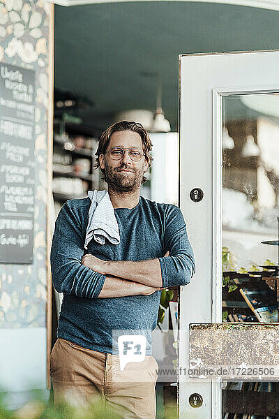 Mature male owner with arms crossed leaning at door at cafe
