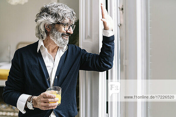 Fröhlicher älterer Mann trinkt Saft und winkt durch das Fenster seines Hauses