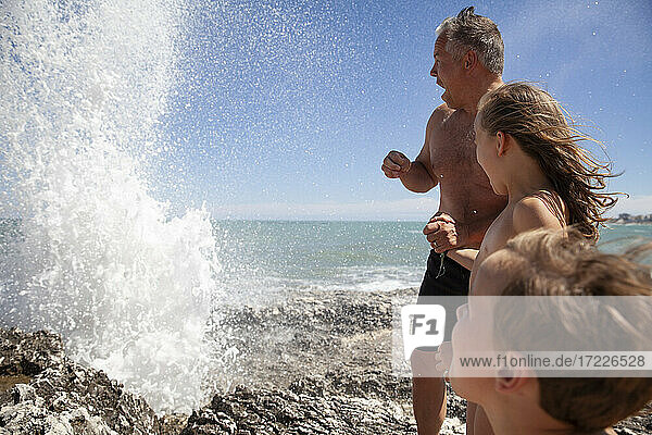 Vater mit Kindern  die auf einem Felsen stehend das Plätschern des Wassers betrachten
