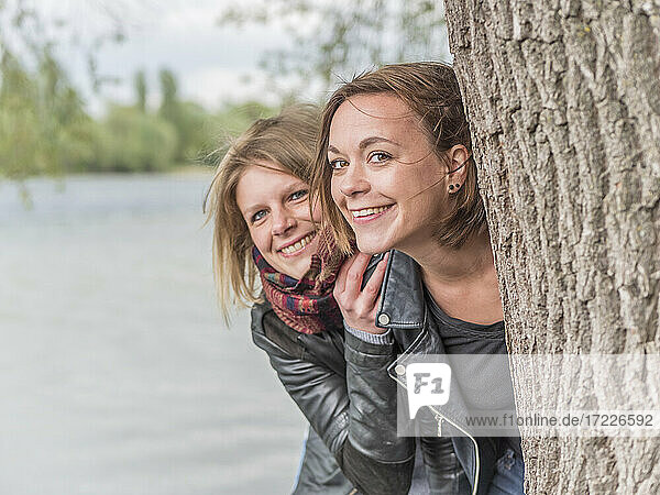 Portrait of two friends hiding behind tree trunk